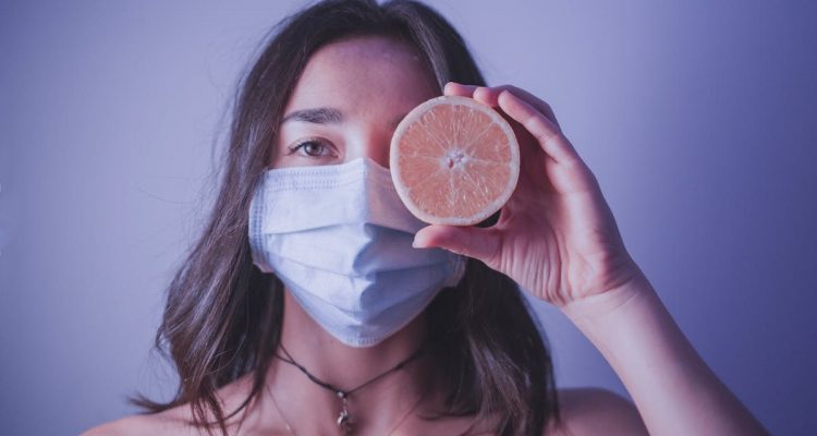 Young female in medical mask looking at camera and covering eye with half of orange while standing near wall during pandemic